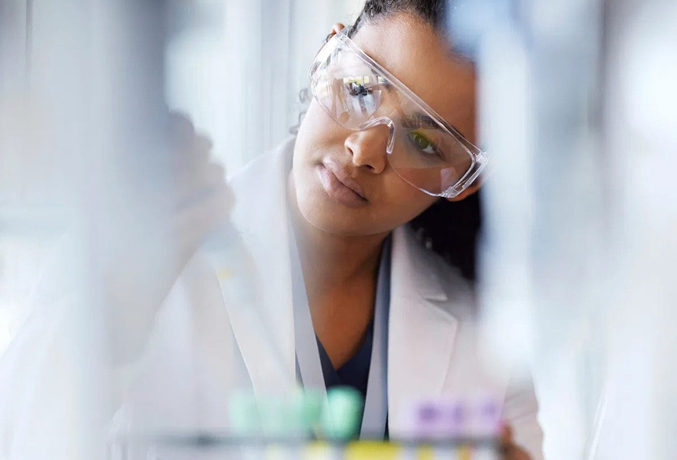 A scientist wearing safety goggles and a lab coat carefully uses a pipette, focusing intently on her work in a laboratory setting with blurred lab equipment in the foreground.