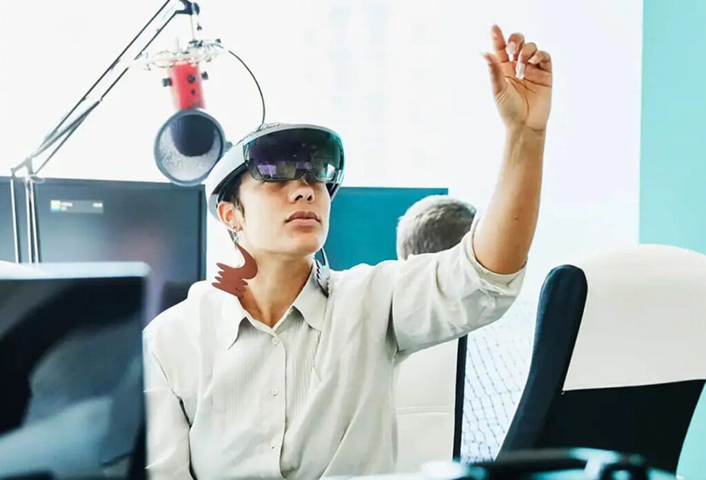 A person wearing augmented reality glasses interacts with the air, appearing to touch a virtual object, while seated at a desk with a microphone and computer equipment in the background.