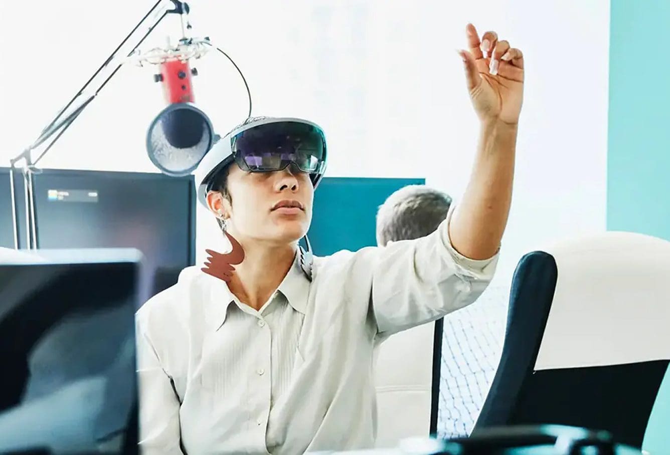 A person wearing augmented reality glasses interacts with the air, appearing to touch a virtual object, while seated at a desk with a microphone and computer equipment in the background.