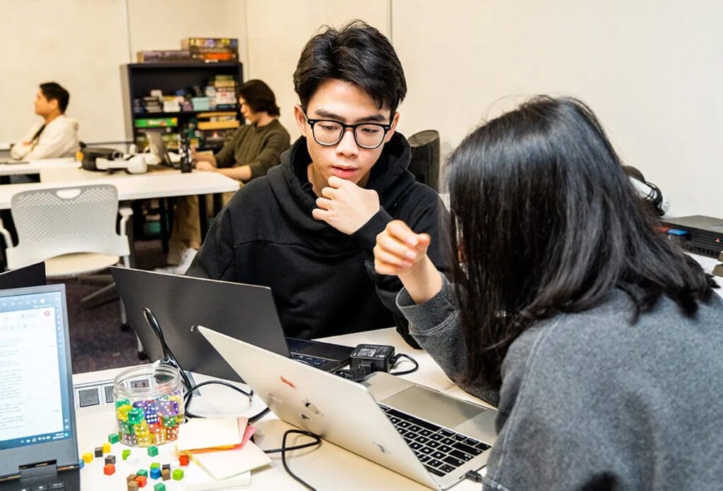Two students sit at a table with laptops, focusing on a task. Colorful dice and notebooks are on the table. More people work in the background near shelves with board games in a bright, modern classroom.
