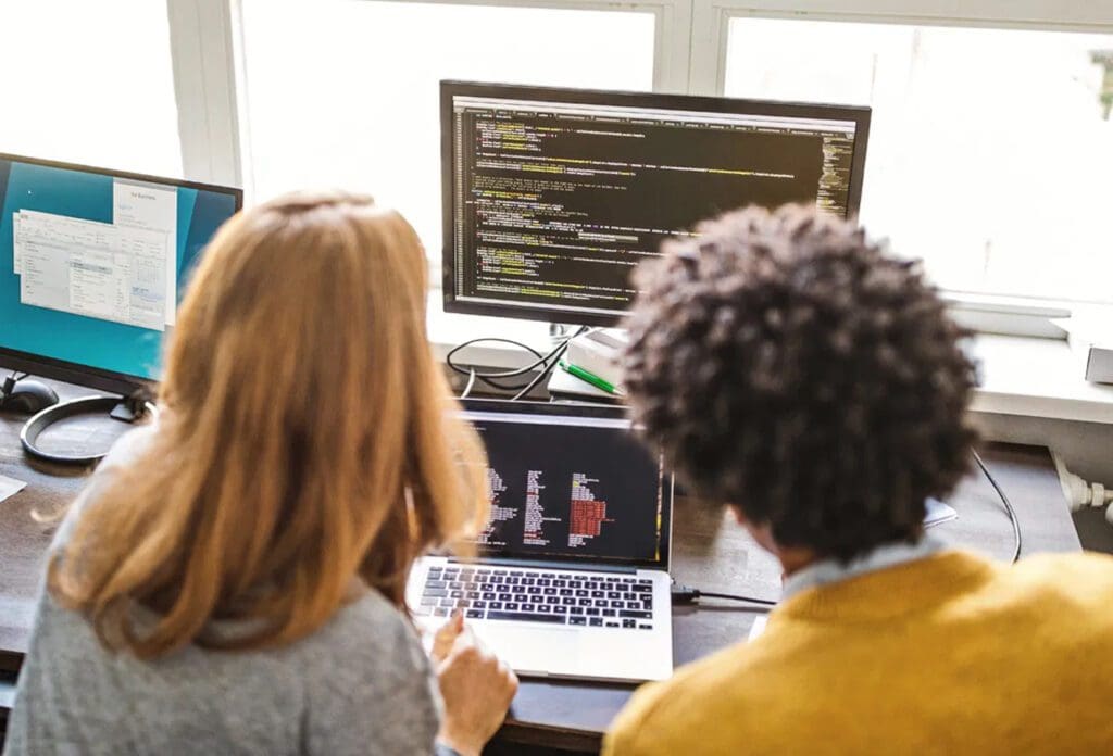 Two people sit at a desk, looking at code on a laptop and a large monitor. Sunlight comes through windows behind them. The scene suggests teamwork or collaborative programming in an office setting.