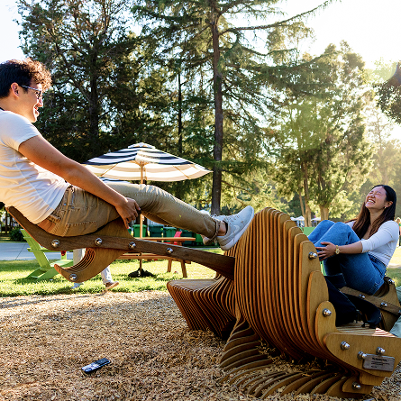 Two people smiling and playing on a wooden seesaw shaped like a fish in a sunny park, surrounded by trees and outdoor seating with a striped umbrella in the background.