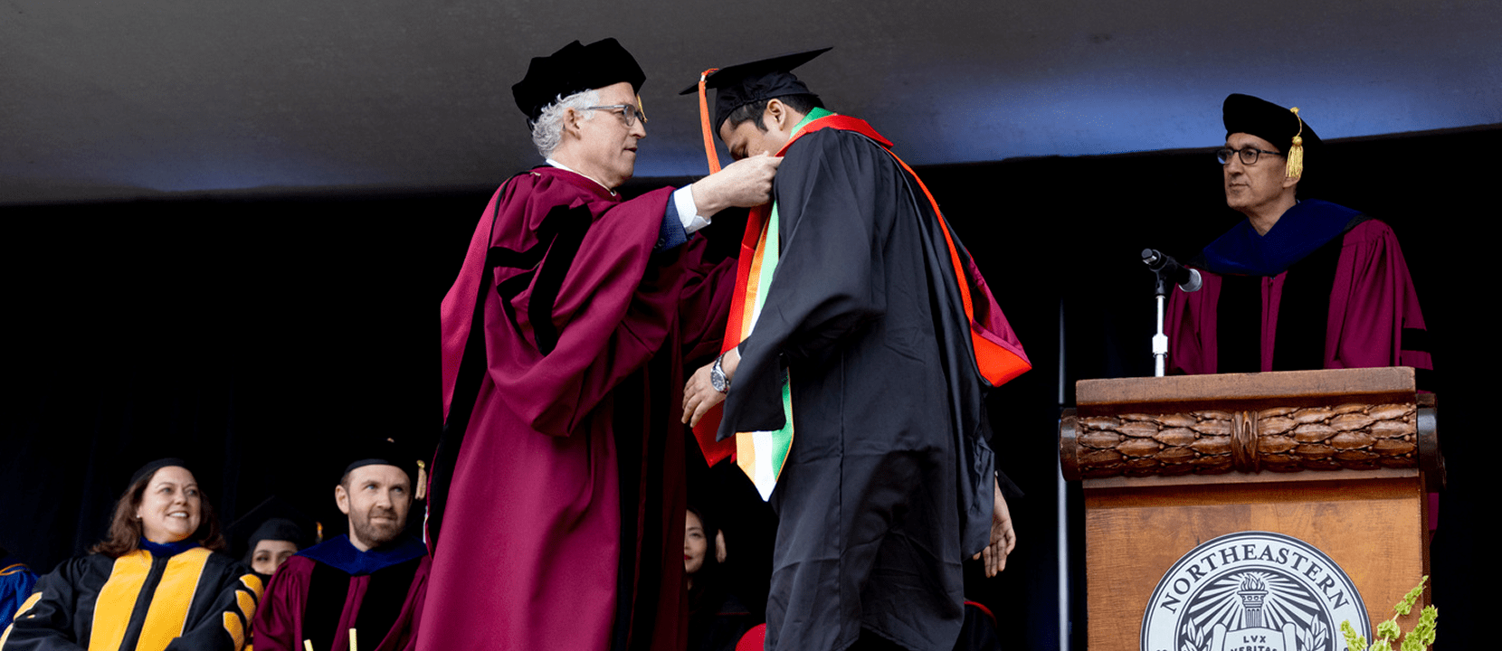 Dean Dan Sachs, wearing a maroon robe, places a graduation hood on a student in cap and gown on stage at a commencement ceremony, with other faculty members seated and standing nearby.