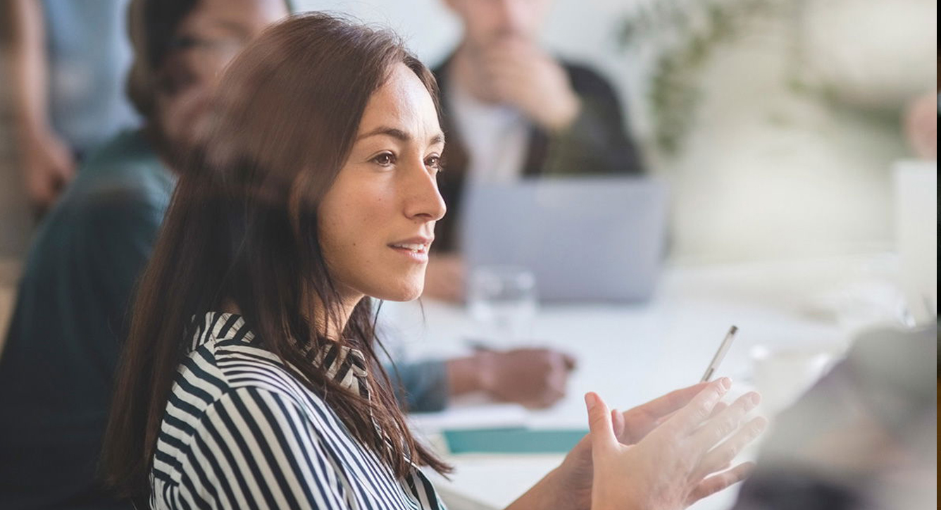 A woman with long brown hair, wearing a striped shirt, speaks and gestures with a pen during a meeting in a bright office, with blurred colleagues and a laptop in the background.