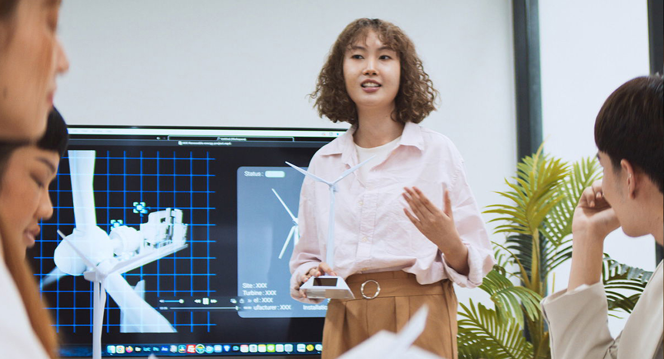 A woman stands and speaks to a group during a presentation, with a large screen behind her displaying technical diagrams of wind turbines and engineering data. Other people sit and listen attentively.