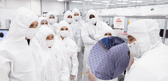 A group of people in white cleanroom suits gathers around a person holding a silicon wafer inside a bright laboratory with shelves and equipment in the background.