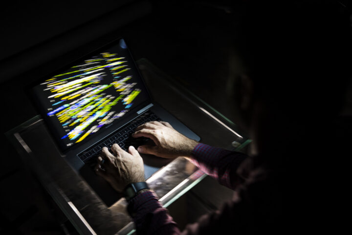 Person typing on a laptop displaying colorful lines of code in a dark setting.