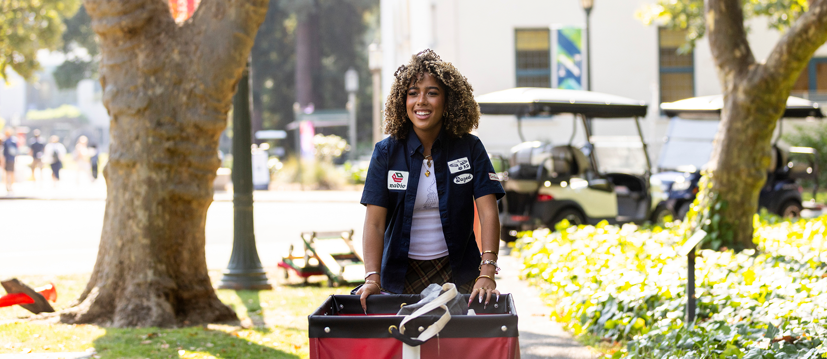 A woman with curly hair, wearing a dark shirt with patches, smiles while pushing a red wagon outdoors on a sunny day. Trees, greenery, and golf carts are visible in the background.