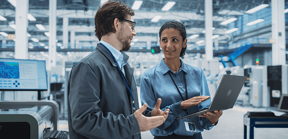 Two professionals in business attire stand in a modern factory, engaged in discussion. One holds a laptop, and both appear to be collaborating. Industrial equipment and machinery are visible in the background.
