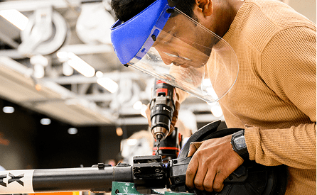 A person wearing a face shield uses a power drill on a metal object in a workshop, focusing intently on their work.