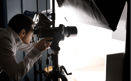 A person operates a professional camera on a tripod in a studio, focusing on a brightly lit setup with softbox lights and photography equipment visible.