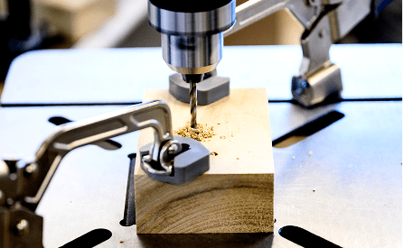 A close-up of a drill press making a hole in a rectangular piece of wood, with clamps securing the wood in place and wood shavings around the drill bit.