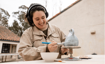 A smiling person wearing headphones and a tan jacket paints a ceramic sculpture shaped like a bird outdoors, using a brush and a bowl of glaze.