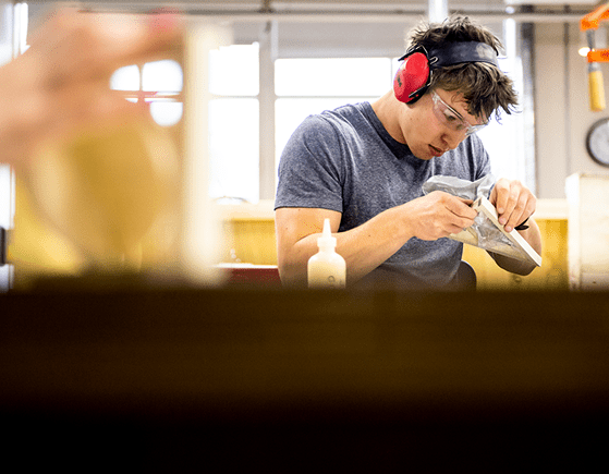 A young man wearing safety goggles and red ear protection carefully works on a woodworking project in a workshop, holding a tool and examining a piece of wood. A bottle of glue sits on the table in front of him.