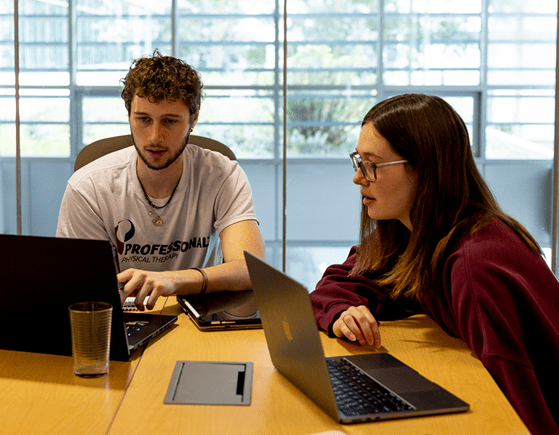 Two students sit at a table with laptops, working together. One wears a white t-shirt, the other a maroon sweater. They appear focused, with a glass of water and a tablet on the table in a bright room.