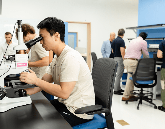 A person sits at a lab table looking into a microscope, while several other people work and talk together in the background of a bright, modern laboratory.