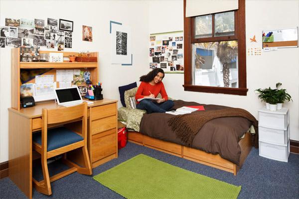 Student sits on bed and writes in room in Orchard Meadow Hall on the Oakland campus of Northeastern University
