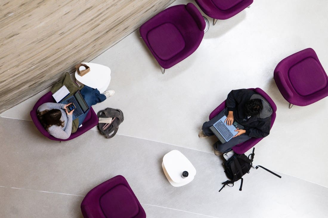 Overhead view of two people sitting apart in purple chairs, each working on a laptop at small white tables. Backpacks are beside them on the light gray floor.