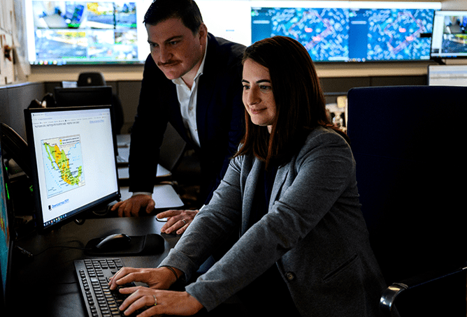 Two people in an office with multiple screens; one is seated and using a computer displaying a map, while the other stands beside her, both focused on the screen. Large monitors with maps and data are visible in the background.
