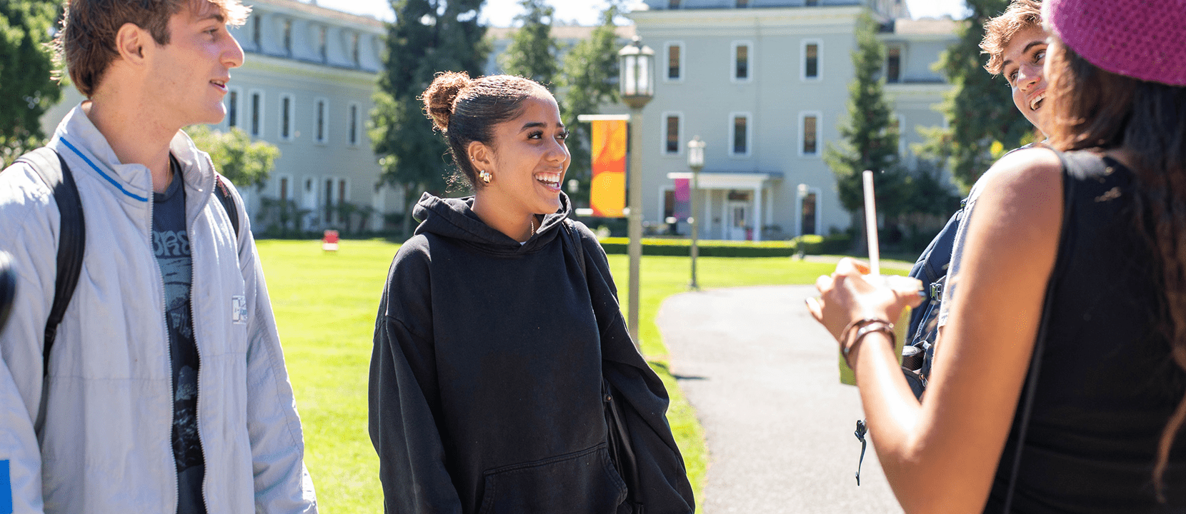 Four students stand outside on a sunny day, smiling and talking on a college campus with grass, trees, and historic buildings in the background.